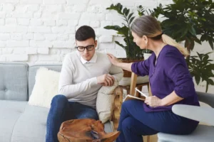 Hombre joven con gafas sentado en un sofá, mostrando signos de preocupación, mientras una mujer de cabello largo y lacio le ofrece apoyo emocional en un entorno acogedor de consulta psicológica.