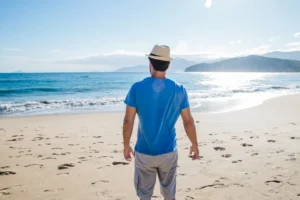 Hombre de pie en la playa, mirando al mar con una camiseta azul y un sombrero de paja, bajo un cielo despejado y soleado. Las olas rompen suavemente en la orilla mientras se aprecian montañas en el horizonte.