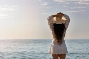 Mujer con cabello rizado y vestido blanco, de espaldas, disfrutando de la vista del mar al atardecer mientras sostiene un sombrero. Ideal para ilustrar temas de vacaciones, verano y bienestar.